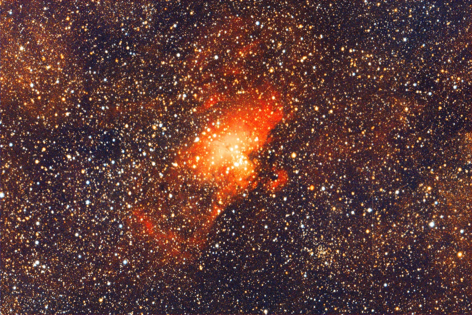 A wide view of the Eagle Nebula as a small red glow within a very dense field of thousands of stars along the Milky Way.
